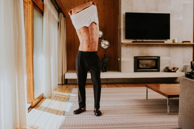 Adult male undressing in a home interior with wood paneled walls, wearing dark pants and sandals, partially removing a white t-shirt.