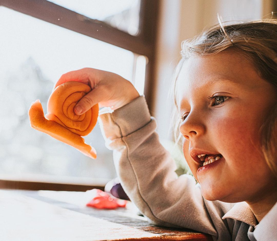 Blond child holds orange playdough snail near window light, showcasing indoor play and handmade toys in a cozy setting.