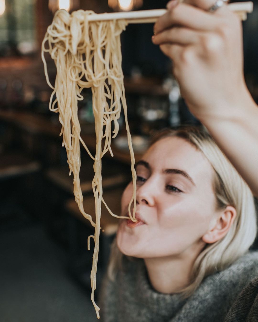 Blonde woman eating spaghetti with chopsticks in an indoor restaurant, showcasing food and beverage consumption.
