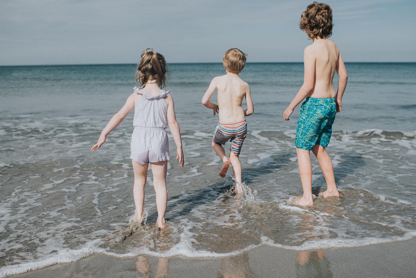 Three children wade in the ocean, backs to camera, on a bright day. One girl in lavender romper, two boys in swim trunks, enjoying the sea.