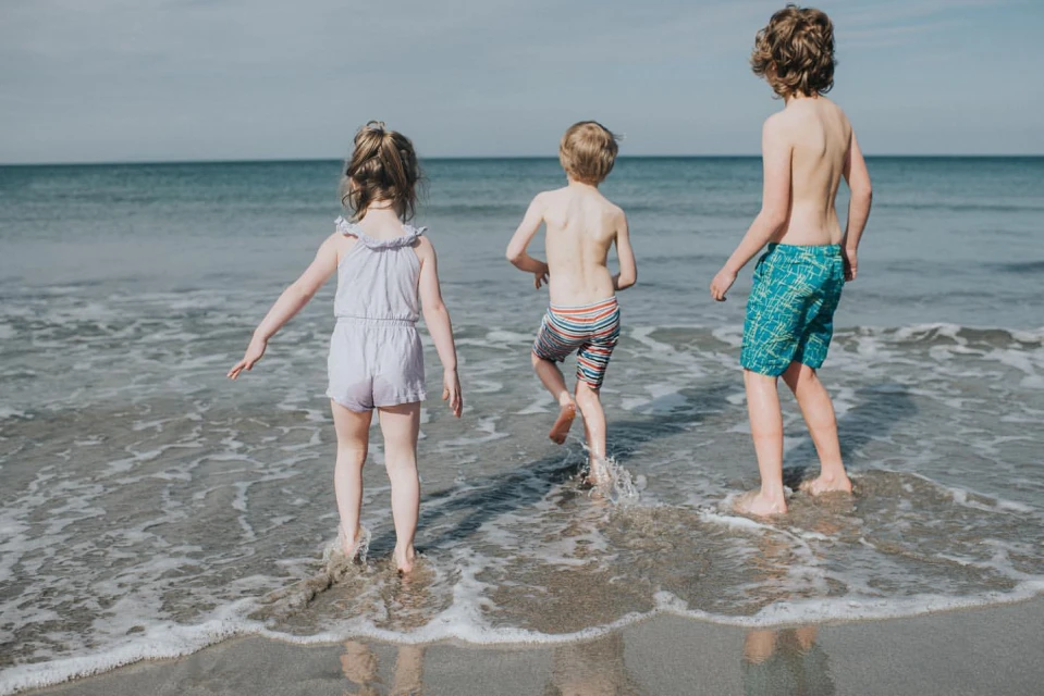 Three children wade in the ocean, backs to camera, on a bright day. One girl in lavender romper, two boys in swim trunks, enjoying the sea.