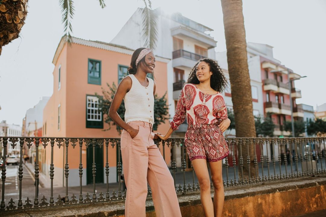 Two women in fashionable summer clothes stand outdoors by a metal railing, smiling and enjoying the warm daylight with palm trees nearby.