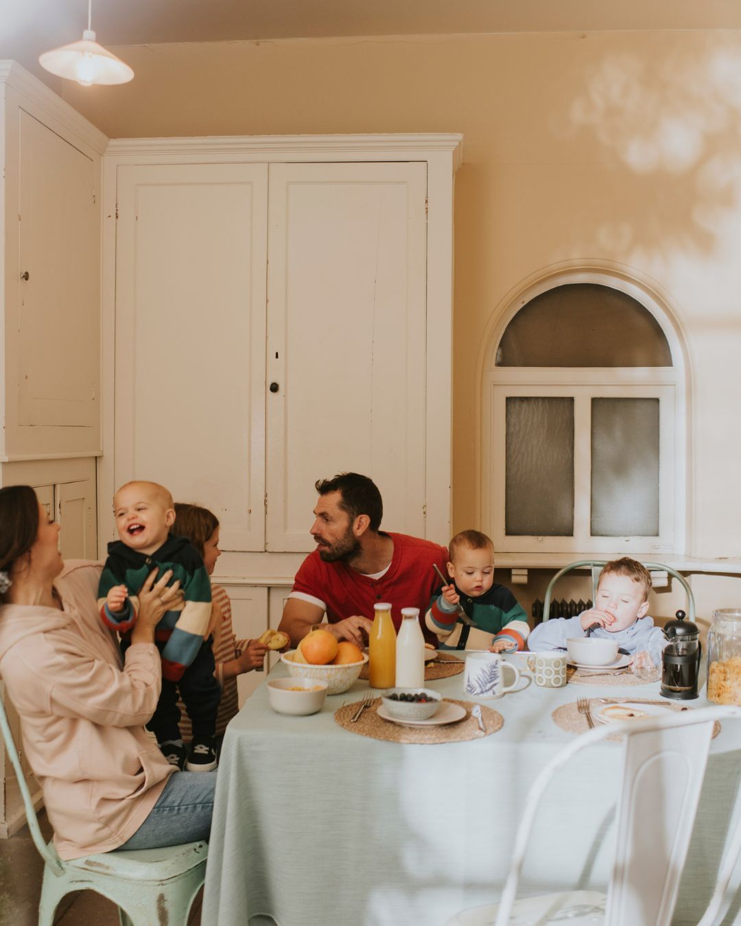 Family gathers at a light-blue clothed table with food, drinks, and dishware for a casual indoor breakfast with light yellow wall.