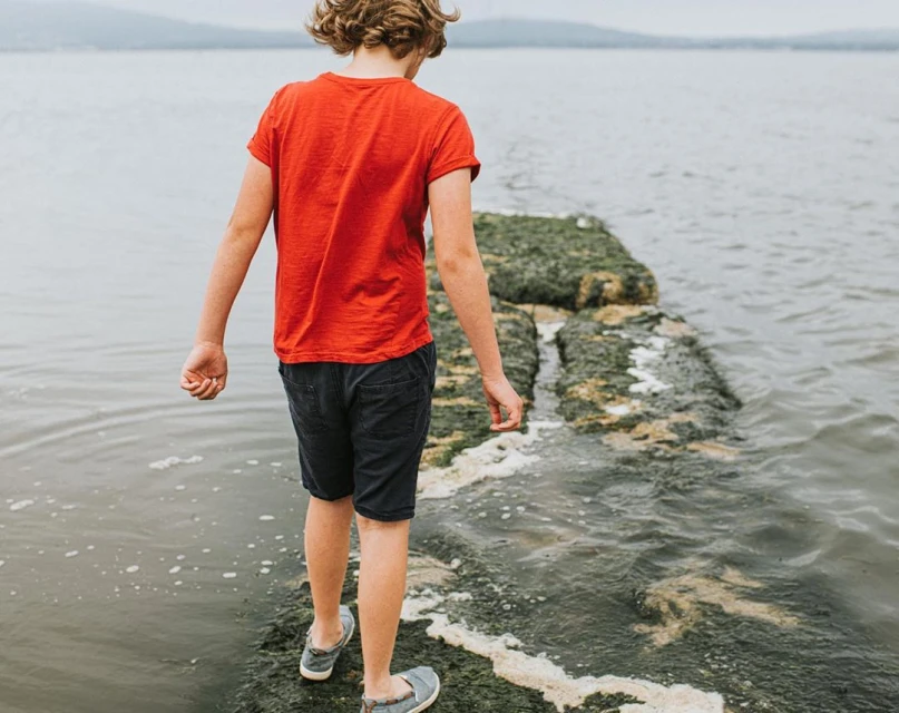 Young child in red shirt walks on concrete path into water on overcast day, landscape background. Youth in dark shorts and shoes.