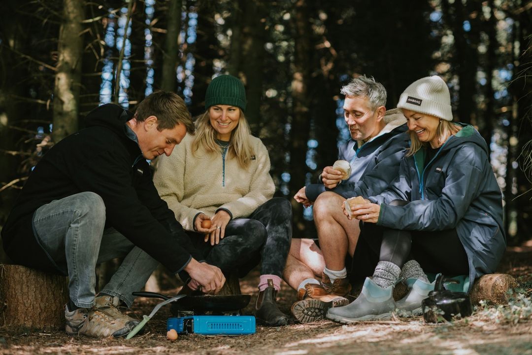 A group of four adults camping in a forest, cooking food on a portable stove, and sitting on logs surrounded by trees.