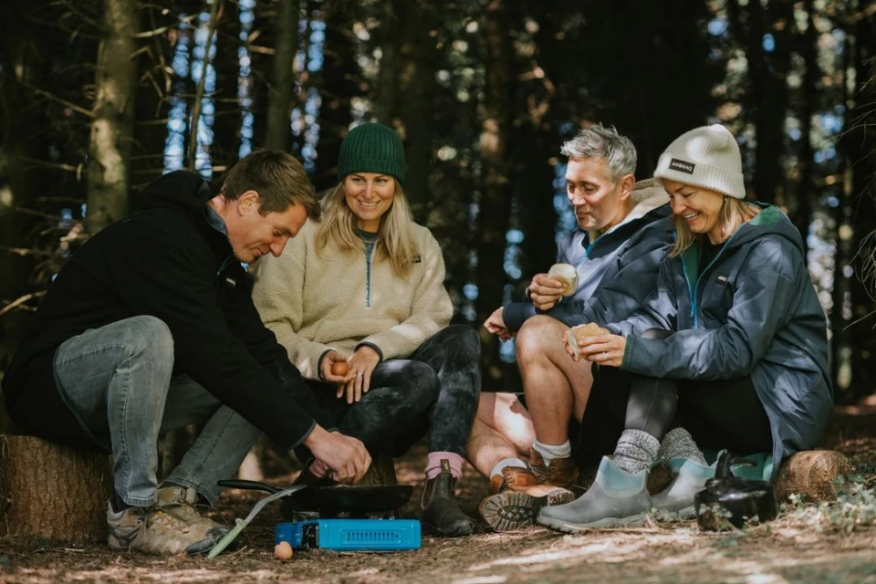 A group of four adults camping in a forest, cooking food on a portable stove, and sitting on logs surrounded by trees.