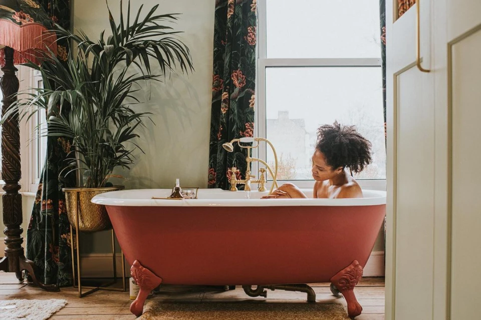 A woman relaxes in a clawfoot bathtub near a window and potted plant, in a room with hardwood floors and floral curtains.