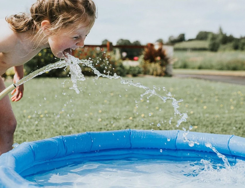 A young child drinks water from a hose, with an inflatable pool in the foreground and a grassy landscape in the background on a sunny day.