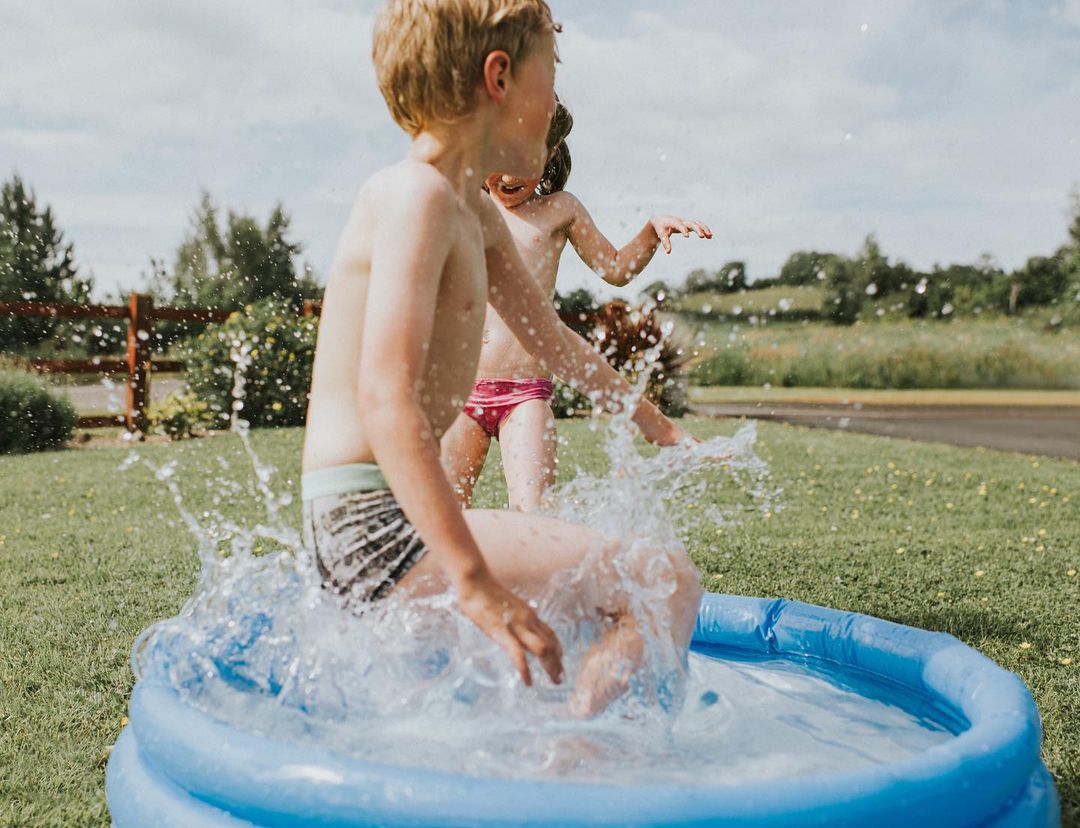 Two children, a boy and a girl, play in a blue inflatable pool on a sunny day, with splashes of water and green grass surroundings.