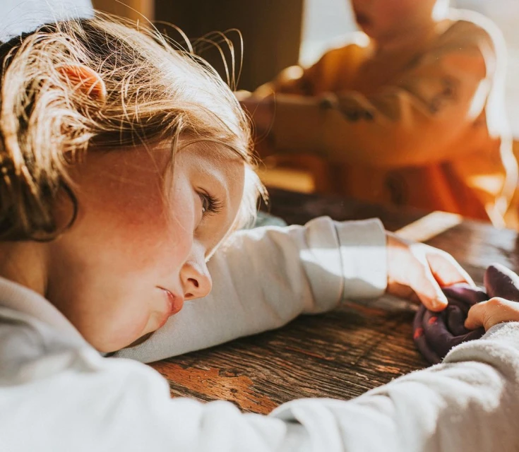 Child rests head on wooden table while playing with modelling clay, with another child visible in the background in a warm, sunlit room.