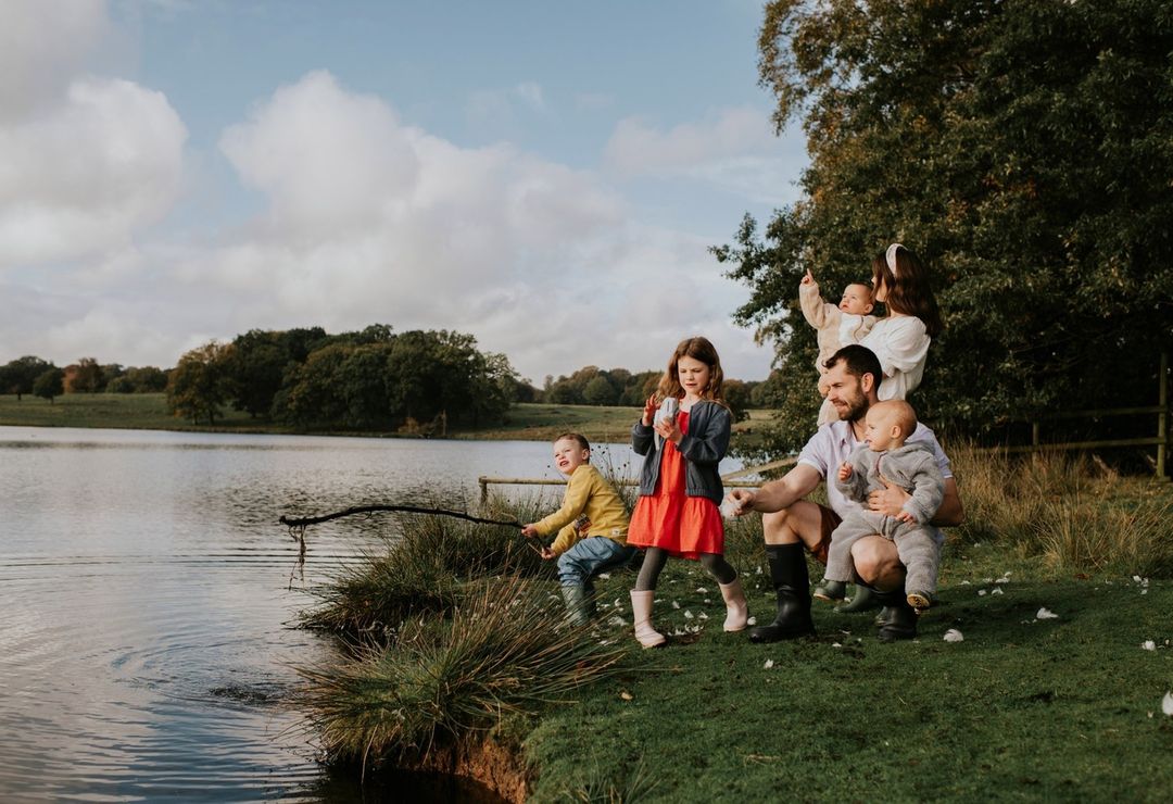 A family of six is gathered near a lake on a cloudy day. A young boy is fishing with a stick, and other family members are nearby.
