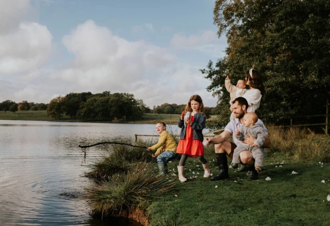 A family of six is gathered near a lake on a cloudy day. A young boy is fishing with a stick, and other family members are nearby.