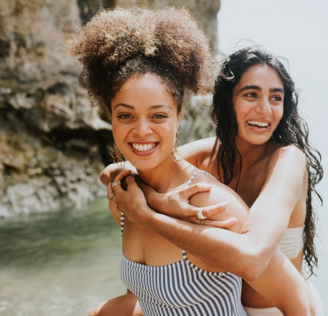 Two young women embrace by the ocean, smiling and wearing swimwear. One has curly hair, and they stand in front of a rocky cliff.
