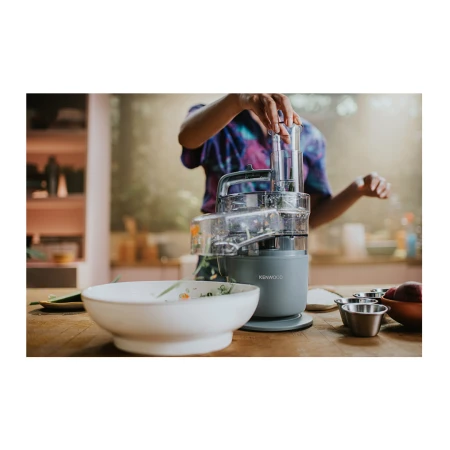 Woman uses a Kenwood food processor on a wood countertop in a bright kitchen, with a white bowl and metal ramekins nearby.