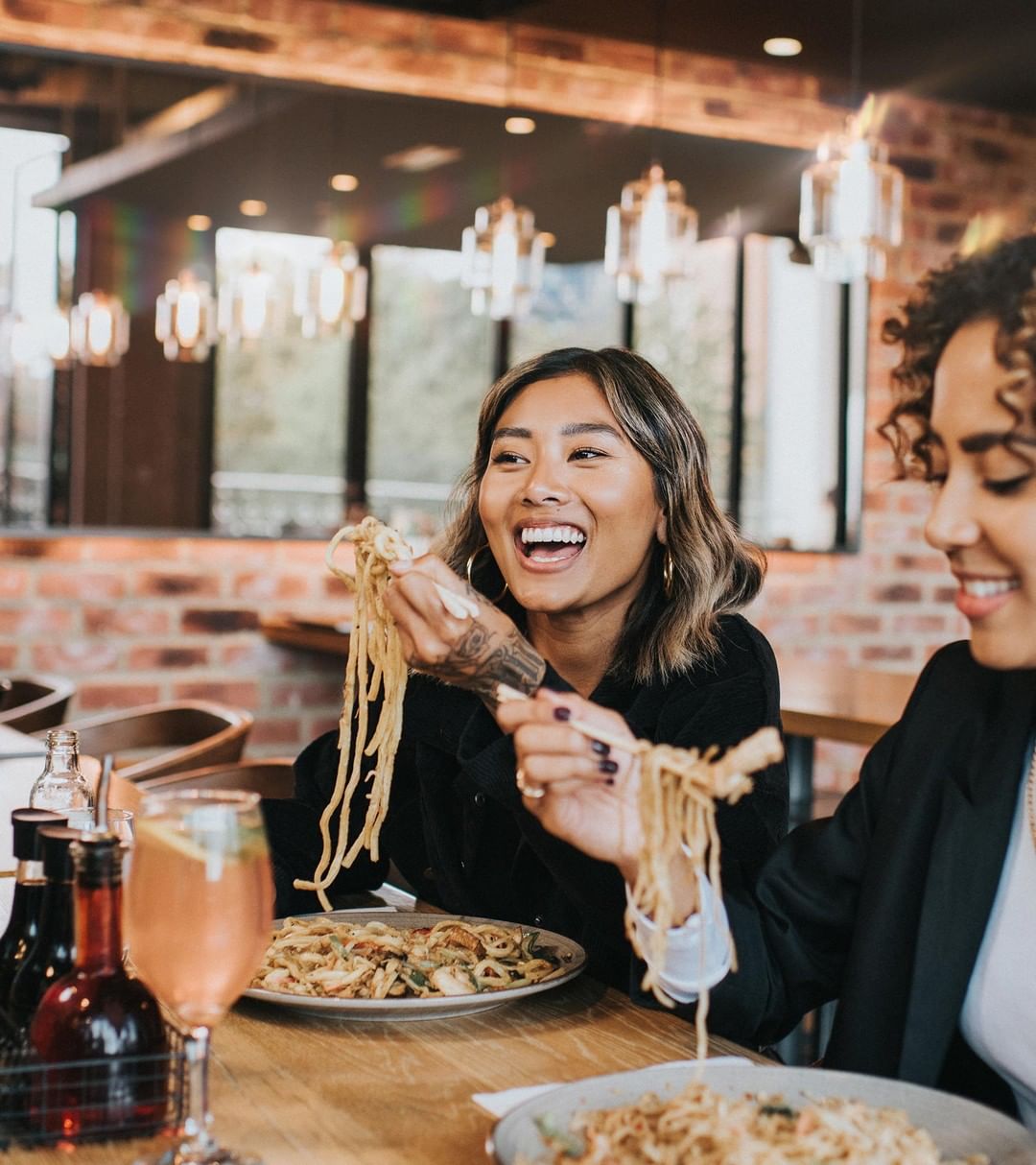 Two women laughing while eating pasta at a restaurant with brick walls and pendant lights, featuring beverages and plates of pasta.
