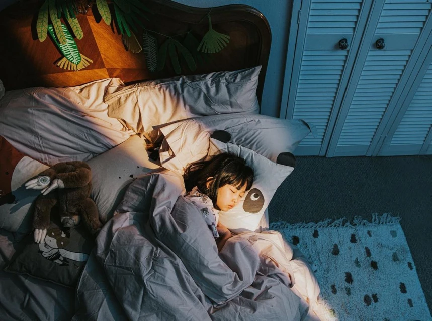 A child sleeps soundly in a bed adorned with leafy decorations, a plush toy, and a panda pillow, under soft, warm lighting.
