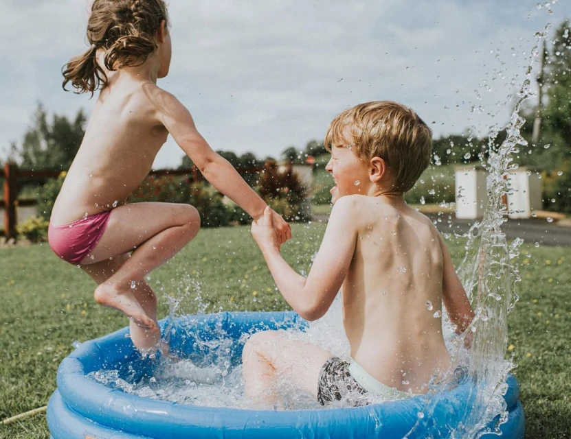 Two children, a girl in mid-air and a boy in the water, play in a blue inflatable pool in a grassy backyard under a bright, cloudy sky.
