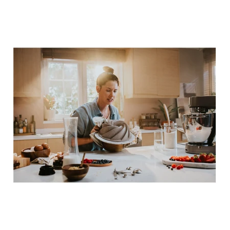 Woman in a kitchen preparing to bake, holding a bundt pan and spritzing it with non-stick spray, stand mixer and other baking supplies visible.