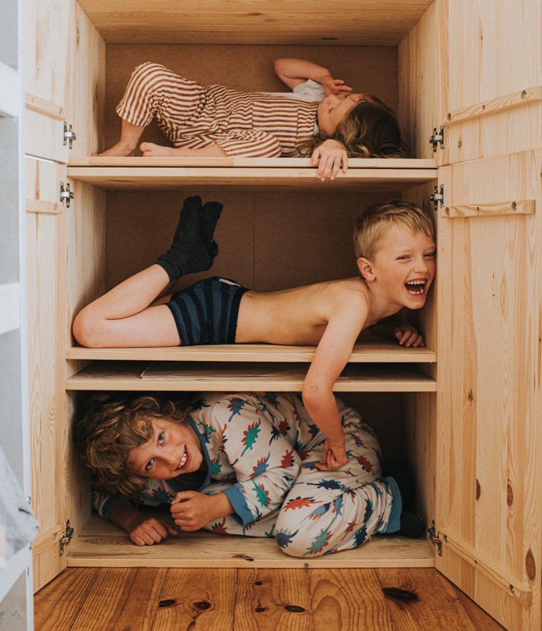 Three children are in a wooden cabinet, each child on a different shelf, smiling and interacting playfully in an indoor setting.