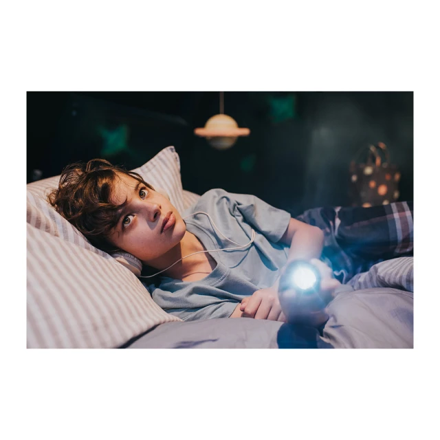 A young boy lying on a bed with headphones, holding a flashlight in a dimly lit room with a planet-shaped lamp. Indoor scene.