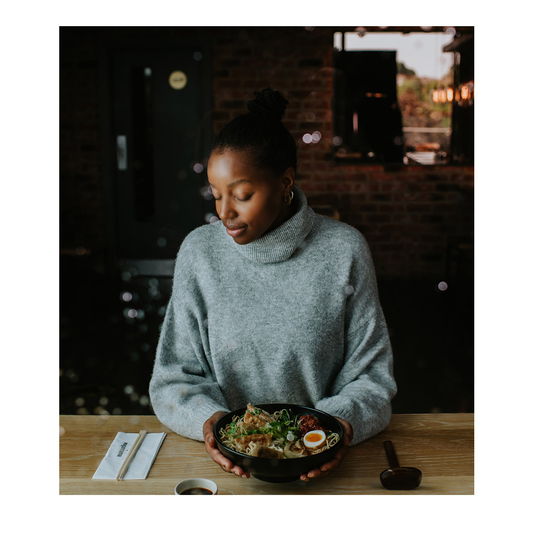 A young Black woman in a gray sweater smiles down at a bowl of ramen she is holding at a restaurant table. Soft, warm light.