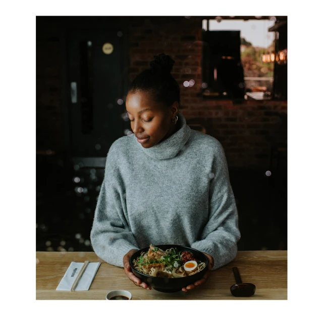 A young Black woman in a gray sweater smiles down at a bowl of ramen she is holding at a restaurant table. Soft, warm light.