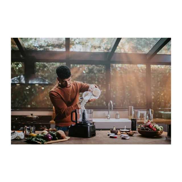 Adult male in an indoor kitchen pouring ingredients from a container into a black food processor, with fruit and vegetables.