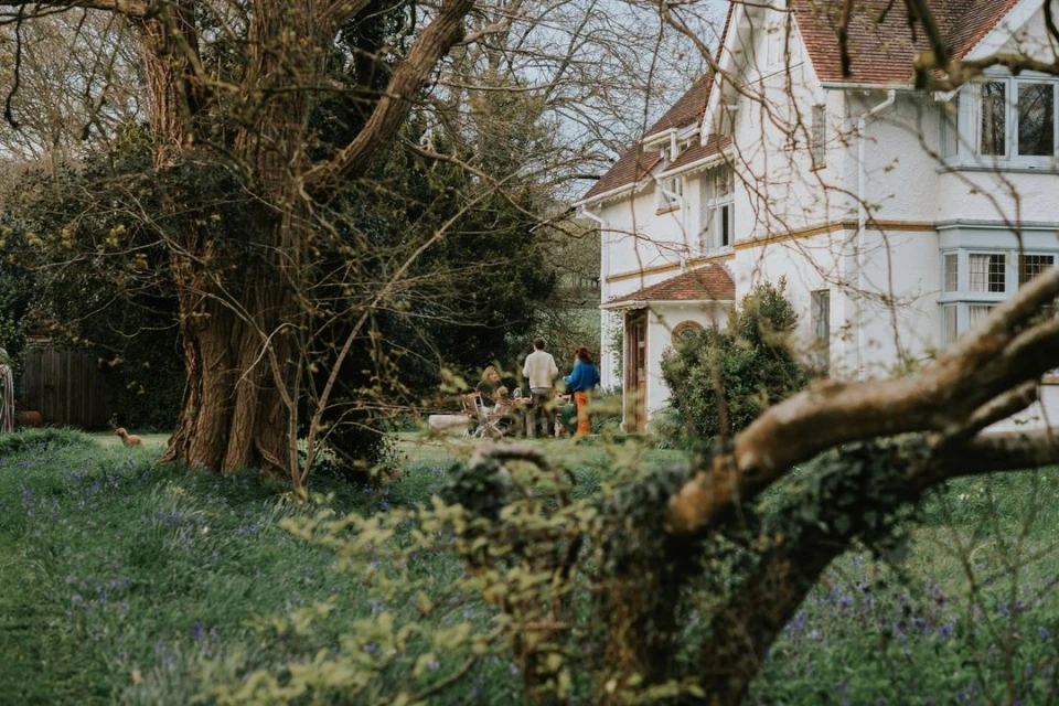 Family gathering outdoors at a long table in the backyard of a large white house surrounded by trees and green grass.