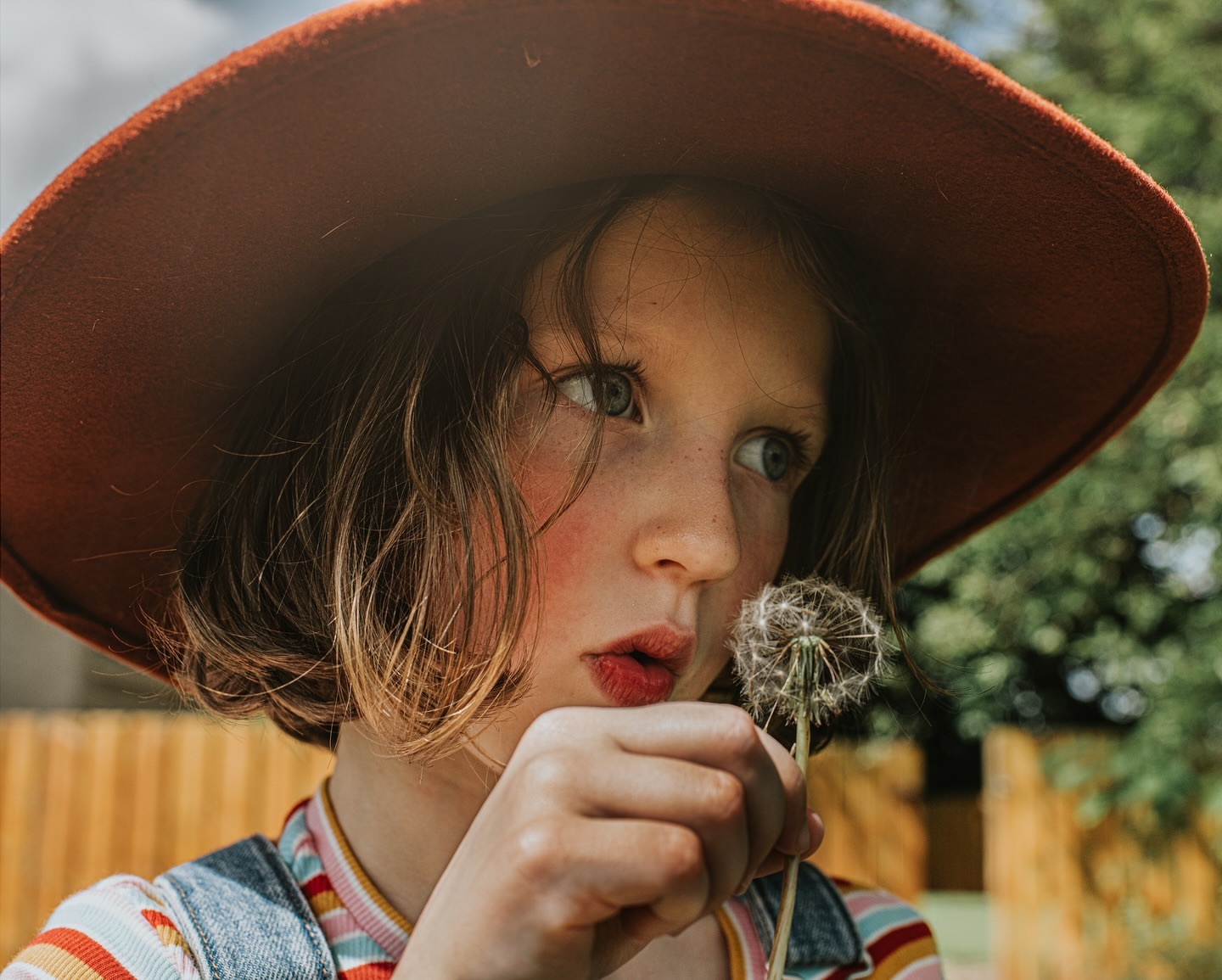 A young girl with a large hat gently blows on a dandelion seed head. She is wearing denim overalls in a natural outdoor setting.