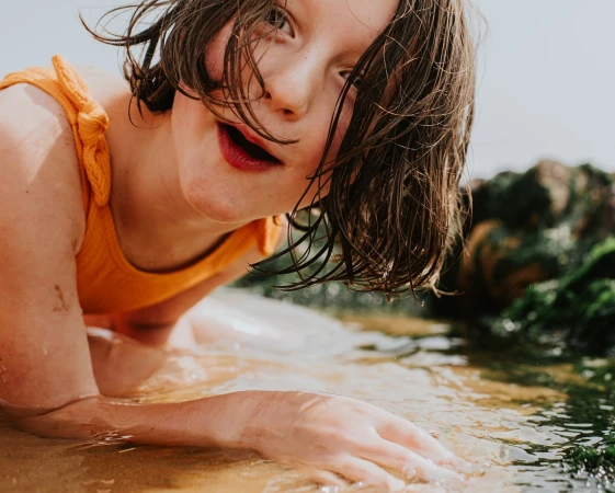 Close-up portrait of a child wearing an orange top leaning over shallow water with wet hair falling across her face.