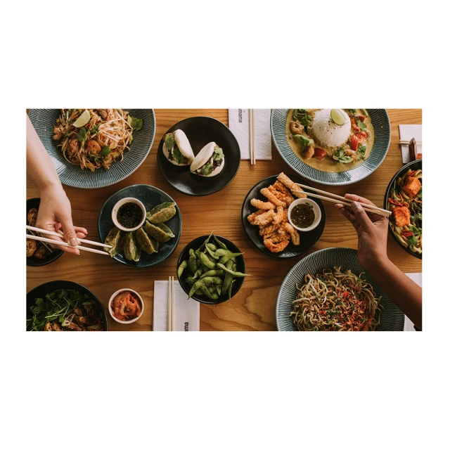Overhead shot of table covered with various Asian-inspired dishes, including noodles, dumplings, buns, and fried food, plus two pairs of hands.