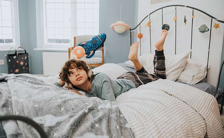 Young boy lies on a bed with a star-patterned blanket and striped bedding, wearing headphones and looking up in an indoor setting.