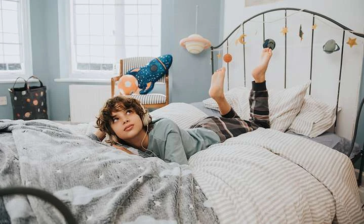 Young boy lies on a bed with a star-patterned blanket and striped bedding, wearing headphones and looking up in an indoor setting.