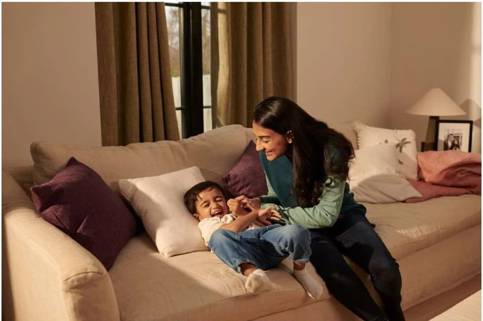 A smiling woman and a child are sitting on a beige sofa in an interior setting with natural light, displaying familial affection and joy.