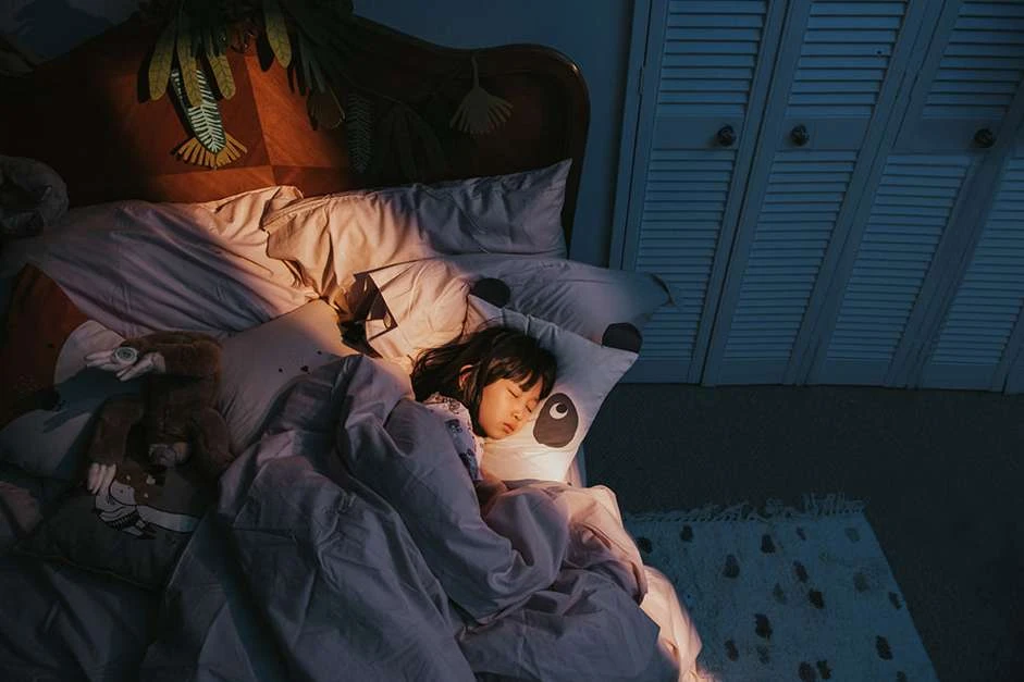 Young child sleeping peacefully in bed with panda pillow and stuffed animal under warm lighting in a cozy bedroom setting.