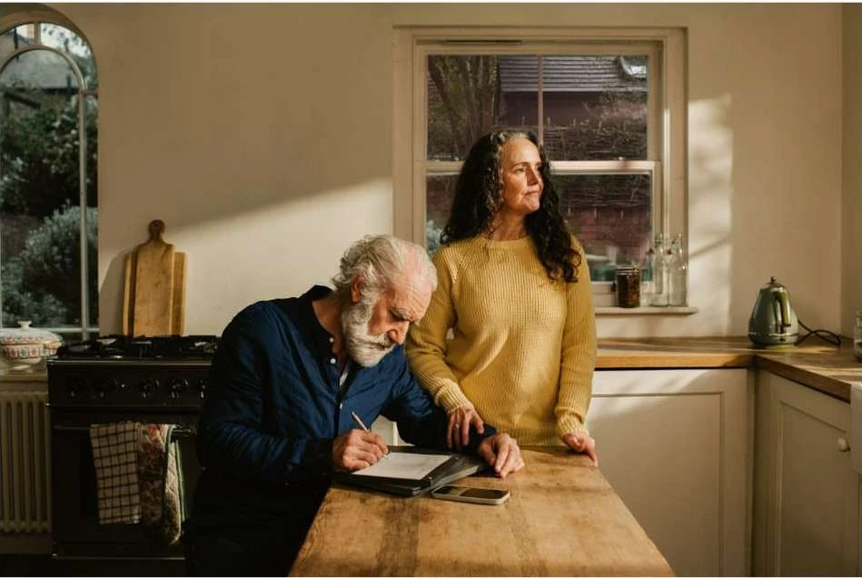 Elderly couple in a warm-toned kitchen; the man writes at a wooden table while the woman gazes out of the window.