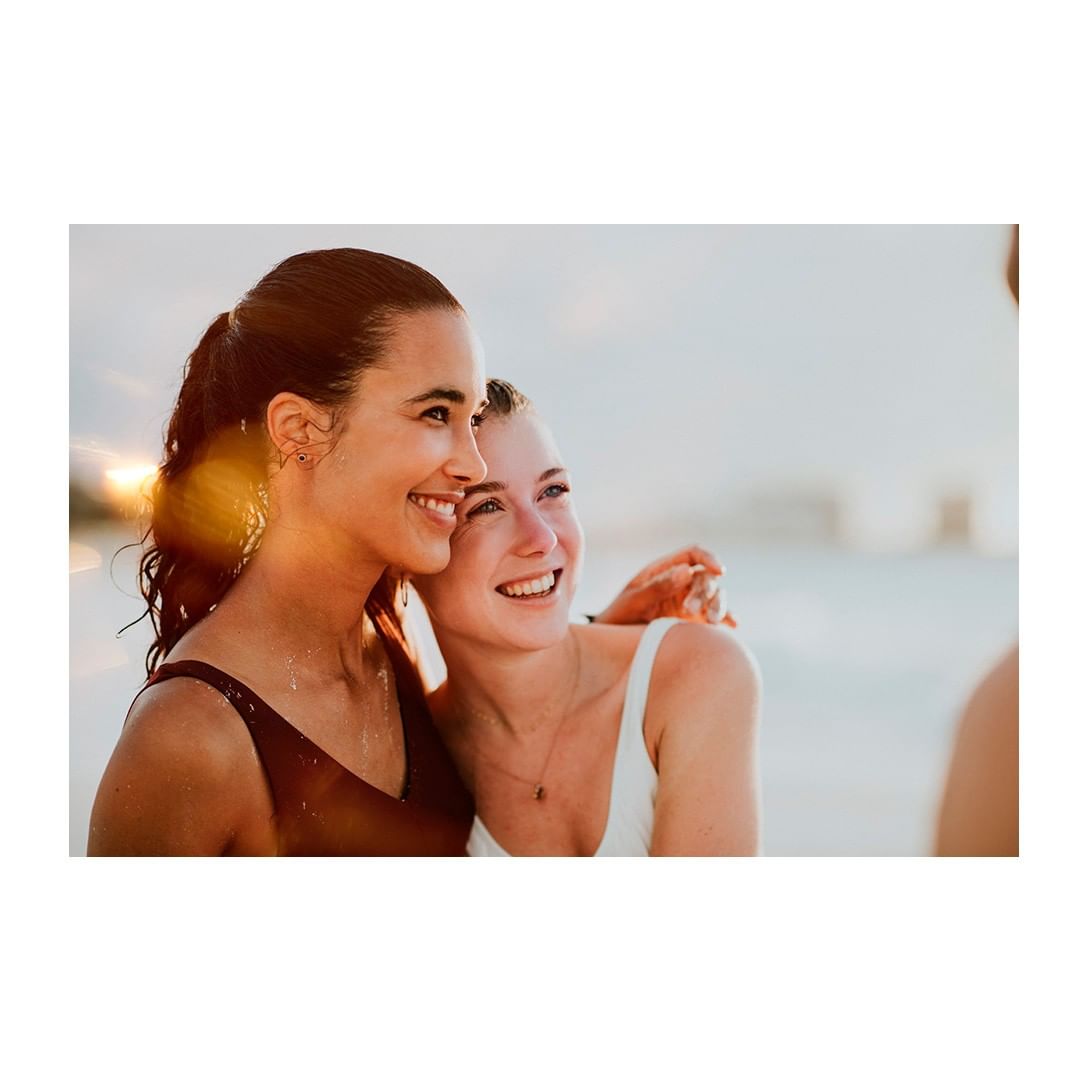 Two smiling women in swimwear embrace on a beach at sunset, creating a warm, friendly atmosphere with sun flare and blurred background.