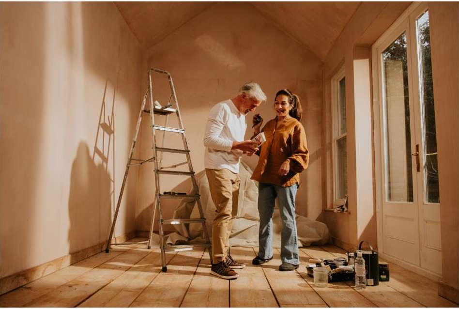 Couple in a room with unfinished walls, woman in jeans and orange jacket, man with grey hair and paint chips, ladder in the background.