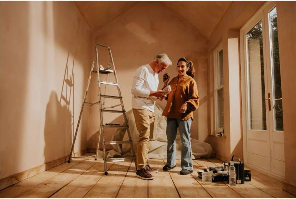 Couple in a room with unfinished walls, woman in jeans and orange jacket, man with grey hair and paint chips, ladder in the background.