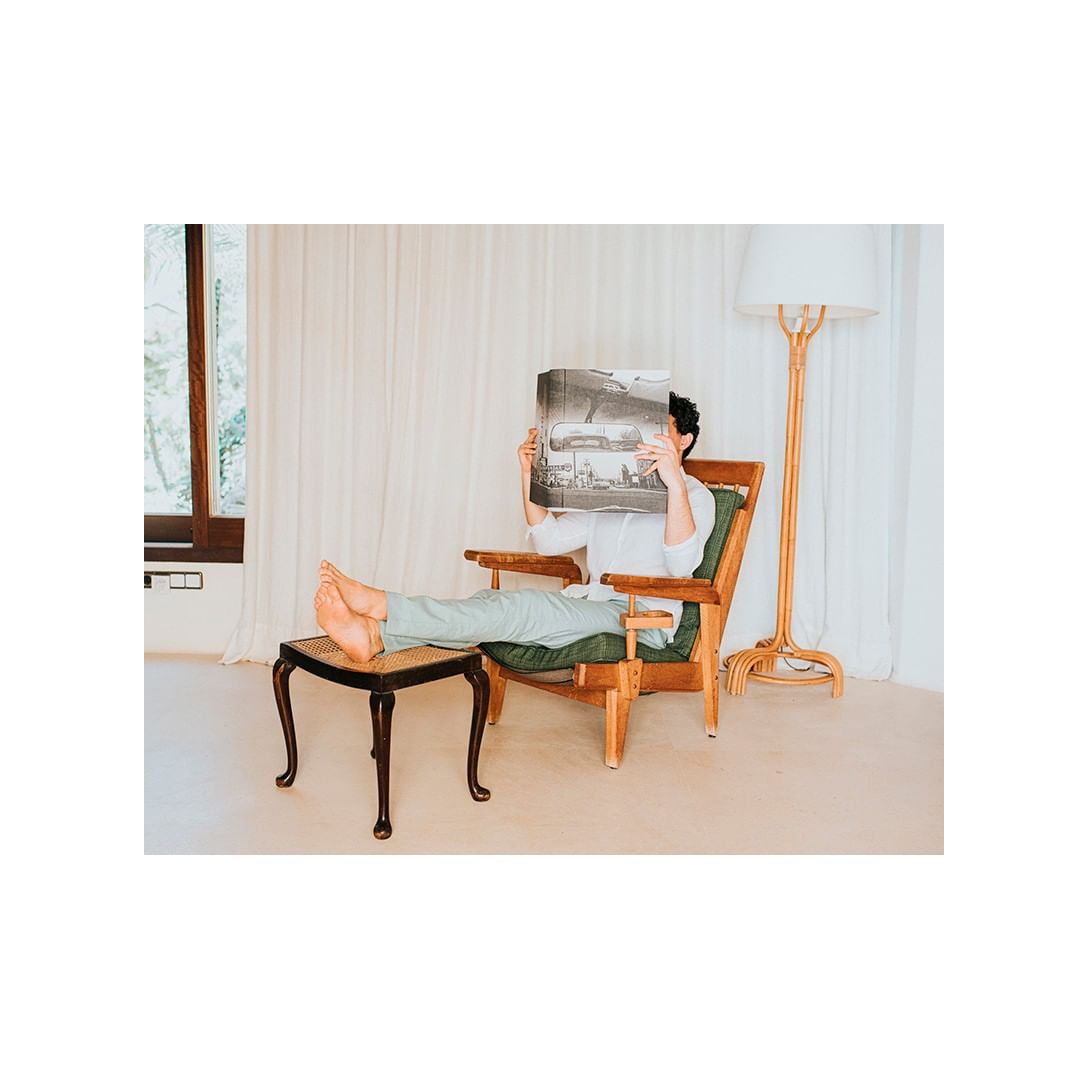 Person relaxing in a wooden armchair, feet up on a stool, reading a book and illuminated by natural light in an indoor setting.