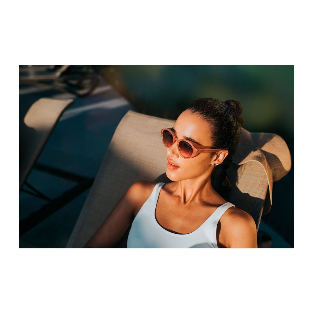 Woman with sunglasses rests on lounge chair outdoors, bathed in sunlight, wearing a white top. Serene and relaxed summer scene.