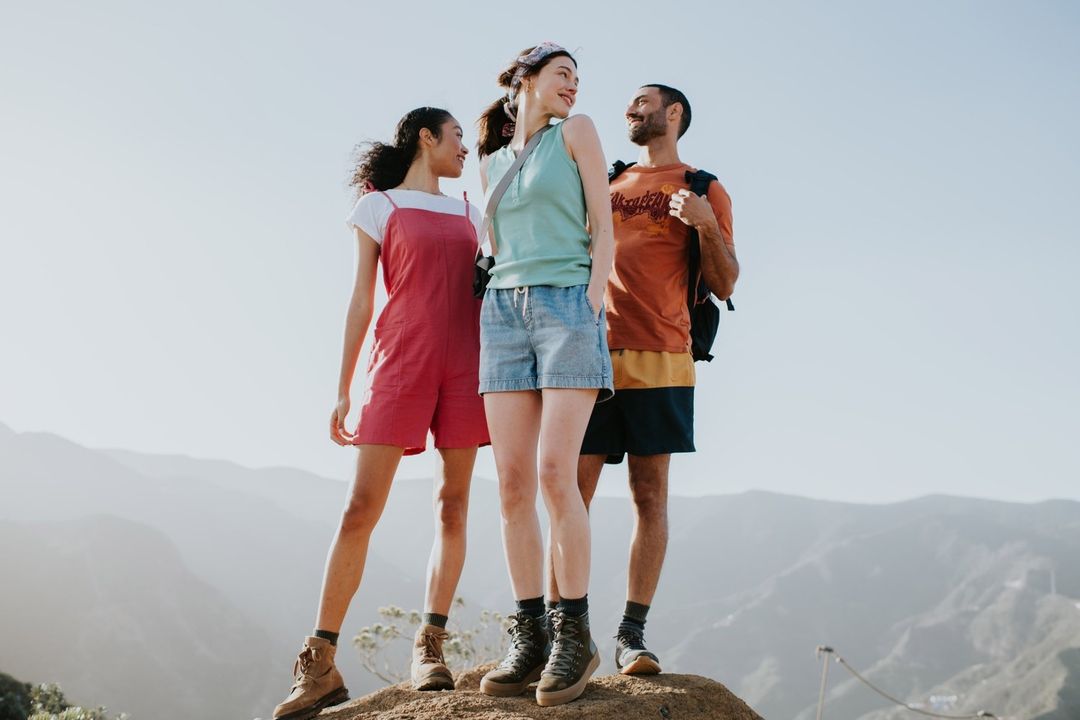 Three friends stand on a mountain top wearing casual hiking clothes. Rolling mountains and clear sky in the background on a sunny day.