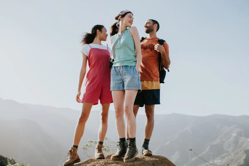 Three friends stand on a mountain top wearing casual hiking clothes. Rolling mountains and clear sky in the background on a sunny day.