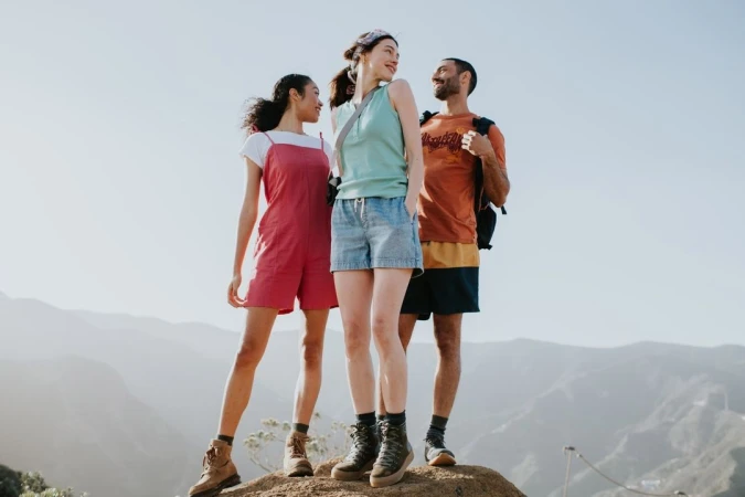 Three friends stand on a mountain top wearing casual hiking clothes. Rolling mountains and clear sky in the background on a sunny day.