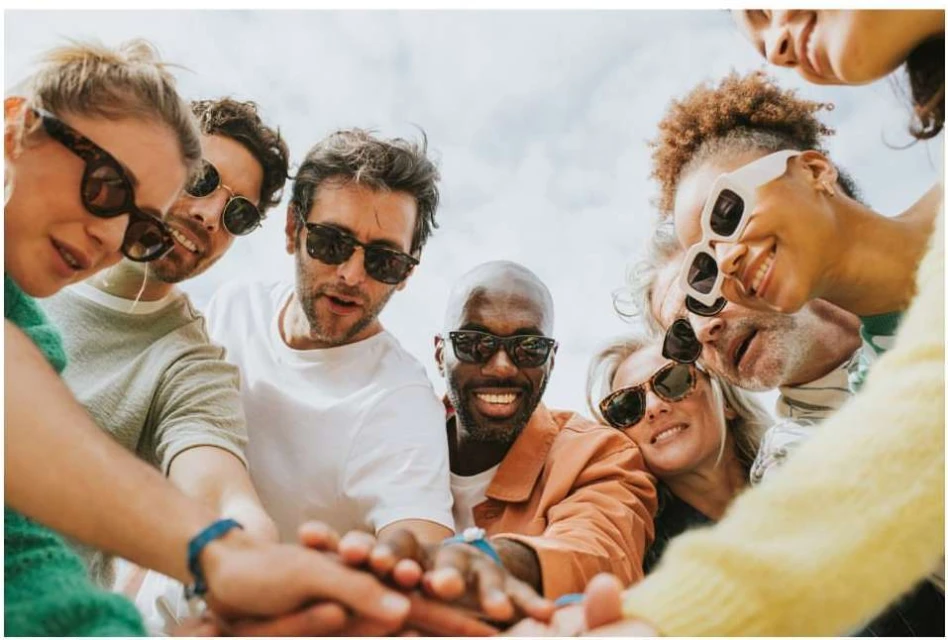 Diverse group of friends smiling, wearing sunglasses, and placing hands together outdoors. Casual and inclusive summer celebration.