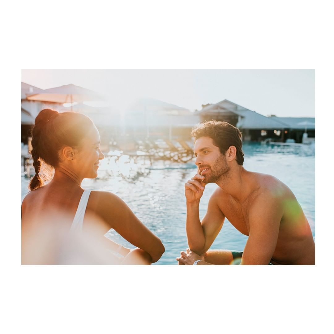 Man and woman in swimwear sit together near a sunlit pool, enjoying a vacation or tropical resort setting, bathed in warm, bright light.