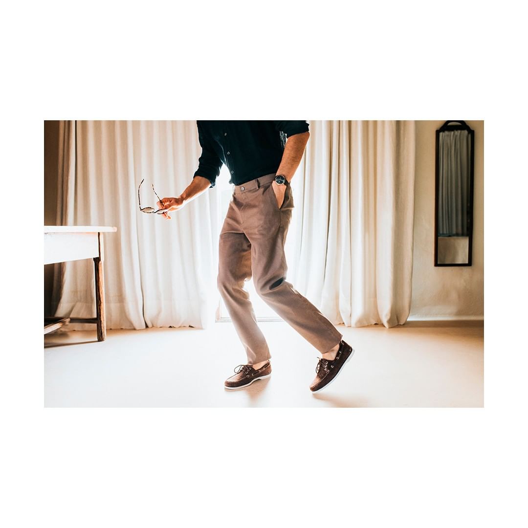 Adult male in a dark shirt and light-brown pants is walking and holding eyeglasses indoors with a cream-colored curtain in the background.