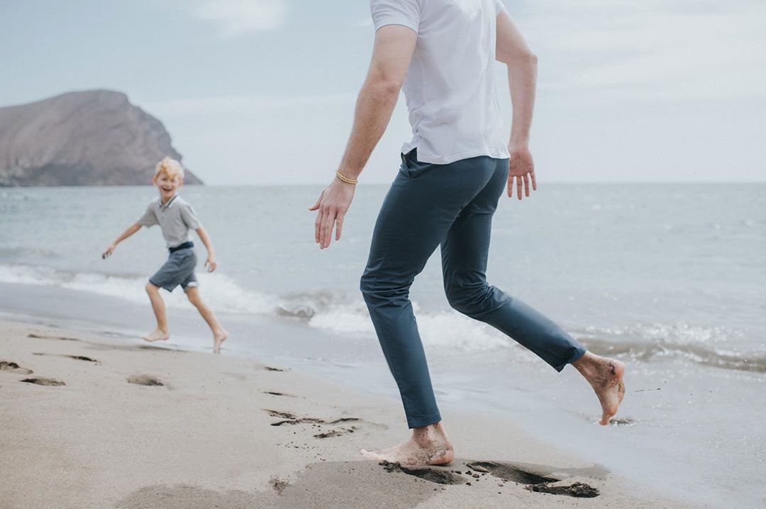A barefoot adult and child are running on the beach near the ocean with a mountain in the background on a sunny day.