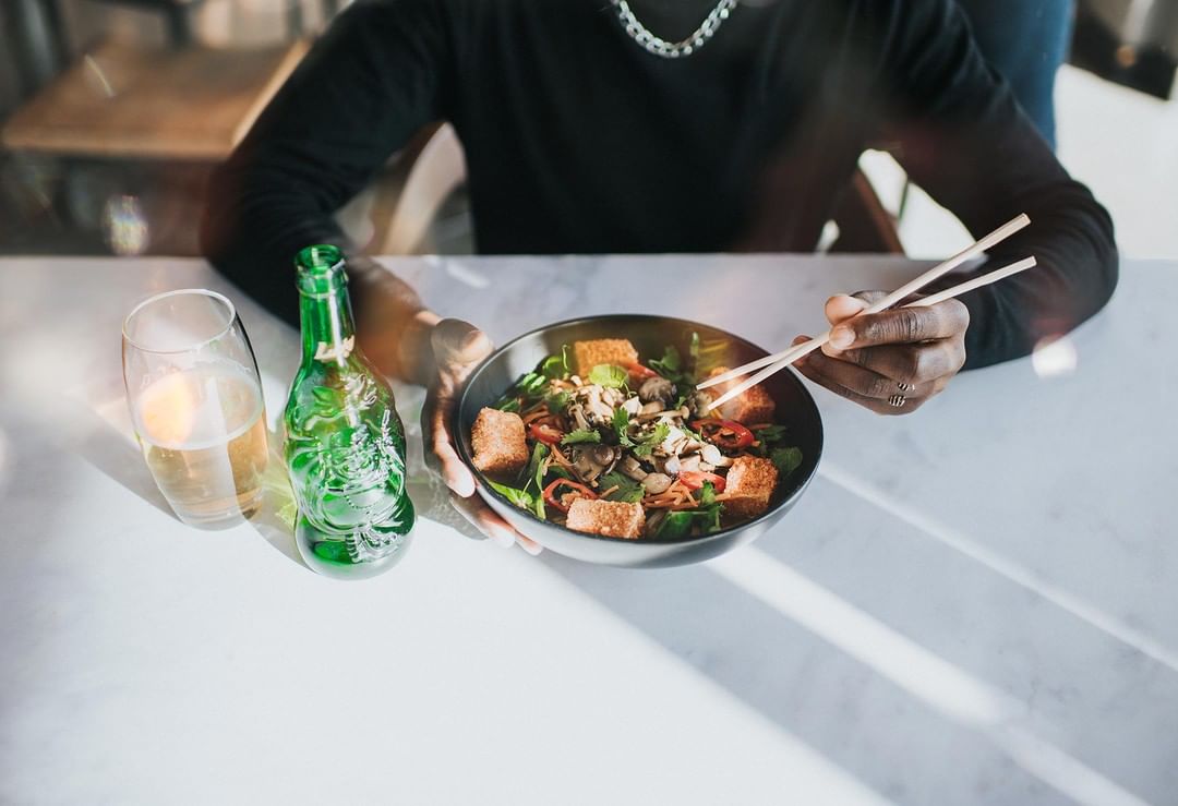 Person eating noodle salad with chopsticks next to a bottle and a glass of beer at a restaurant table with natural light.