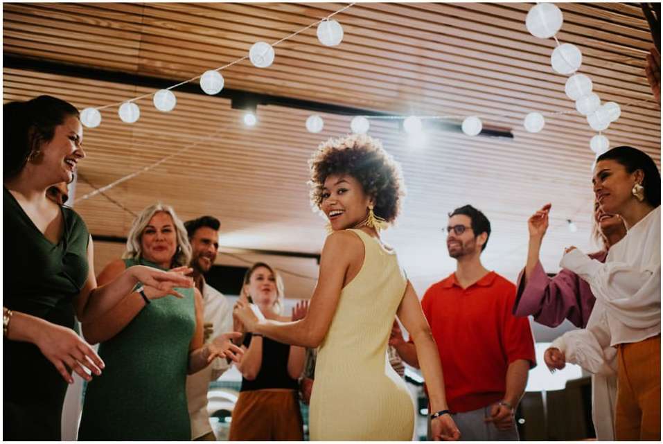 A group of eight people are gathered together indoors, smiling and dancing under white string lights in a joyful, candid setting.